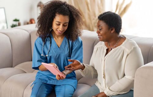 Elderly dark-skinned lady feeling joyful while talking to nurse Talking to nurse. Elderly dark-skinned lady feeling joyful while talking to nurse showing her news on tablet